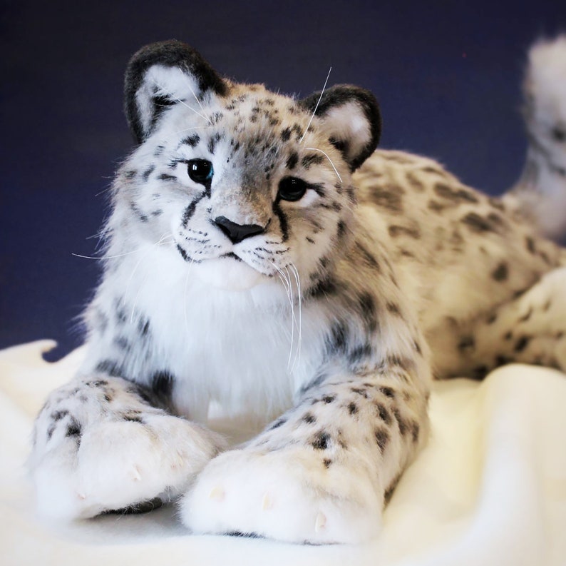 May include: A white and gray plush snow leopard with blue eyes. The snow leopard is lying on a white surface with its paws outstretched.