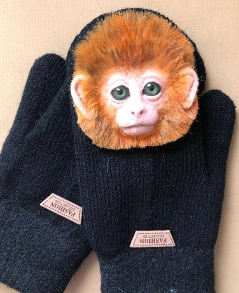 Puede incluir: Mitones de punto negro con un parche de cuero marr&oacute;n que dice "Fashion Collection". Una peque&ntilde;a cara de mono realista est&aacute; unida a la parte superior de los mitones.