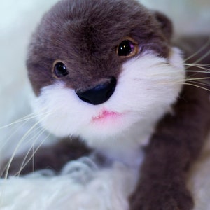 Op de afbeelding: Een pluche otter met grijs haar, wit haar rond zijn neus en mond, en zwarte ogen. De otter ligt op een wit, pluche oppervlak.
