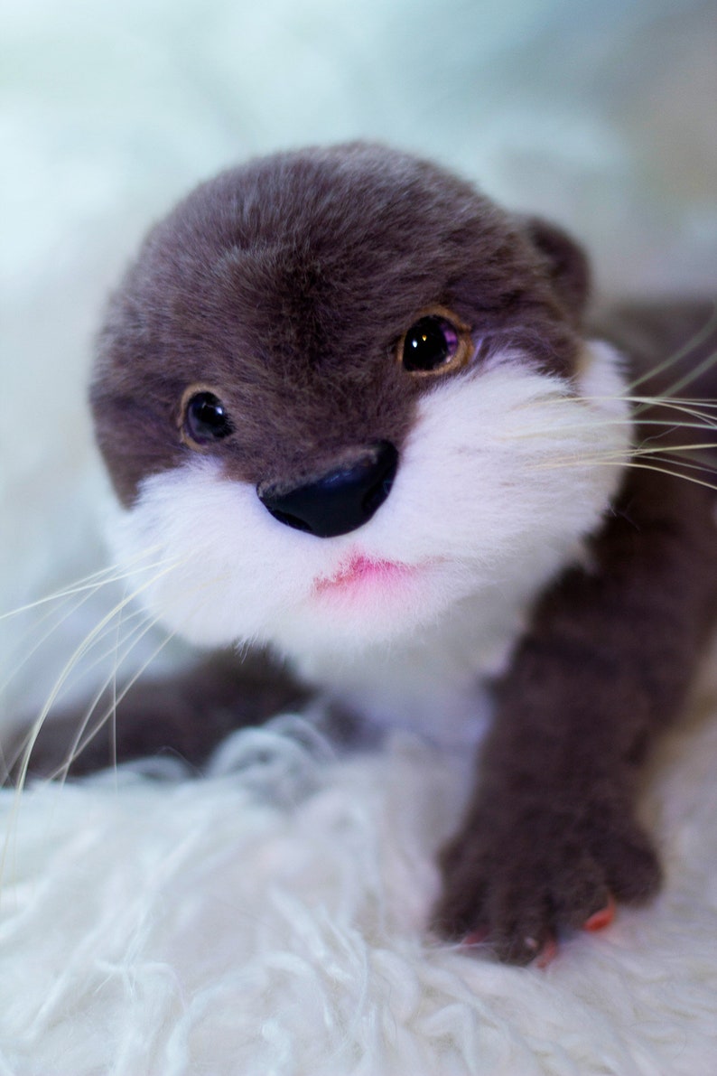 Op de afbeelding: Een pluche otter met grijs haar, wit haar rond zijn neus en mond, en zwarte ogen. De otter ligt op een wit, pluche oppervlak.