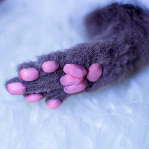 Op de afbeelding: Close-up van een grijze kattenpoot met roze voetkussentjes. De poot rust op een wit, pluche oppervlak.