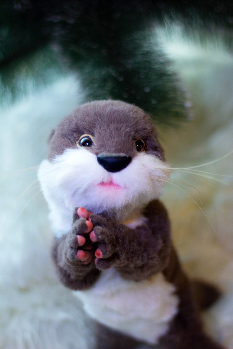 Op de afbeelding: Een grijze en witte pluche otter met roze klauwen, zittend op een wit, pluche oppervlak. De otter heeft een lieve uitdrukking en houdt zijn pootjes bij elkaar.
