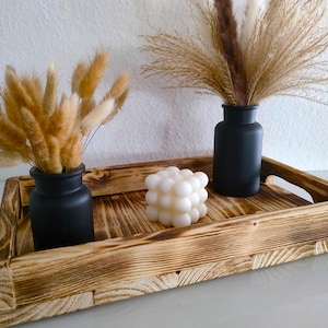 Könnte beinhalten: Ein rustikales Holztablett mit Griffen, das zwei schwarze Vasen mit Trockenblumen und eine weiße Blasenkerze trägt. Das Tablett hat eine natürliche Holzmaserung und eine gebrannte Oberfläche, was eine warme Ästhetik erzeugt.