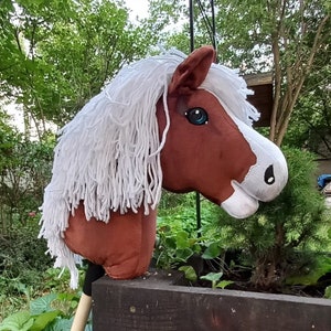 May include: A brown and white plush horse head on a stick, with a long white mane. The horse has blue eyes and is mounted on a wooden post. The toy is designed for children and is set against a backdrop of greenery.