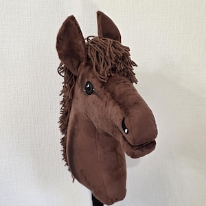 May include: A brown plush hobby horse toy featuring a soft fabric head with upright ears, dark brown yarn mane, and embroidered eye and nostril details. The horse head is mounted on a cylindrical pole, showcasing a realistic equine shape.