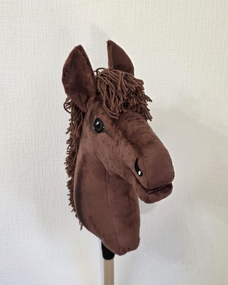 May include: A brown plush hobby horse toy featuring a soft fabric head with upright ears, dark brown yarn mane, and embroidered eye and nostril details. The horse head is mounted on a cylindrical pole, showcasing a realistic equine shape.