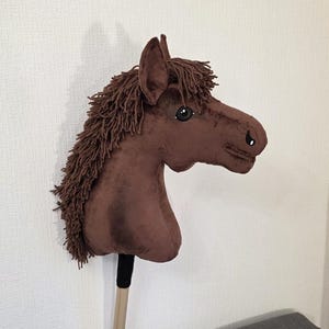 May include: A brown plush hobby horse with a soft fabric head, dark brown yarn mane, and black and white eyes. The horse head is attached to a light-colored wooden stick with a black fabric wrap at the base of the neck, creating a toy horse.