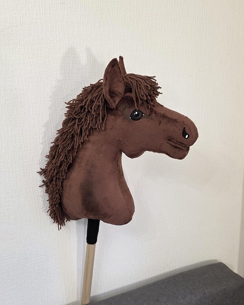 May include: A brown plush hobby horse with a soft fabric head, dark brown yarn mane, and black and white eyes. The horse head is attached to a light-colored wooden stick with a black fabric wrap at the base of the neck, creating a toy horse.