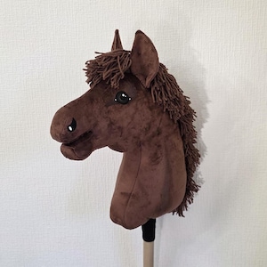 May include: A brown plush hobby horse toy featuring a soft fabric head with dark brown yarn mane and forelock. The horse has embroidered eyes and nostrils, pointed ears, and is mounted on a smooth, light-colored wooden stick handle.
