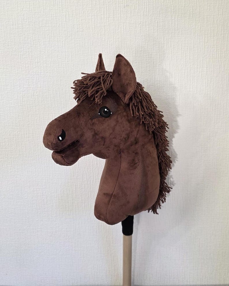 May include: A brown plush hobby horse toy featuring a soft fabric head with dark brown yarn mane and forelock. The horse has embroidered eyes and nostrils, pointed ears, and is mounted on a smooth, light-colored wooden stick handle.