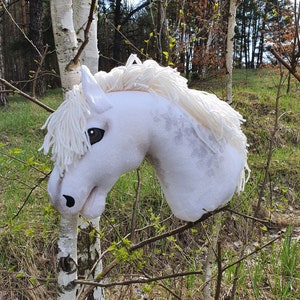 Puede incluir: Cabeza de caballo de peluche blanca con una crin y cola blancas. La cabeza del caballo está parcialmente oculta por un tronco de abedul. La cabeza del caballo está en un entorno boscoso.