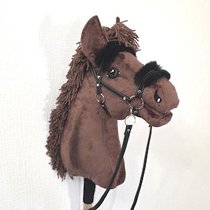 May include: A brown plush hobby horse toy featuring a dark brown yarn mane, black faux leather bridle with silver-toned metal rings, and black faux fur trim on the noseband and browband, all mounted on a wooden stick handle for play.