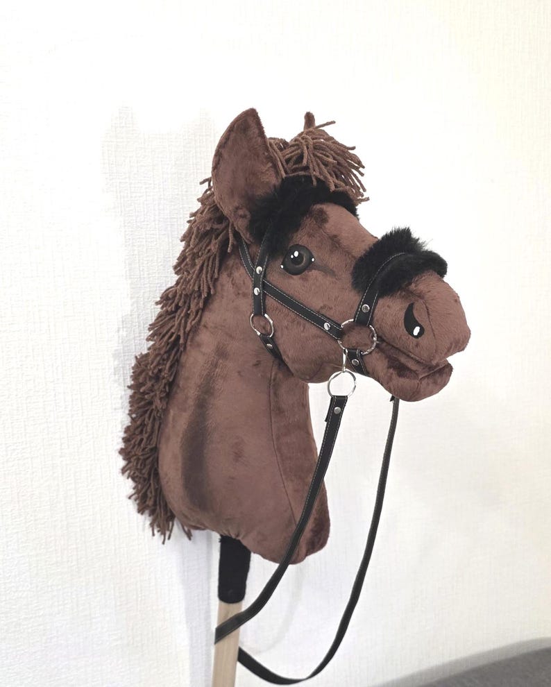 May include: A brown plush hobby horse toy featuring a dark brown yarn mane, black faux leather bridle with silver-toned metal rings, and black faux fur trim on the noseband and browband, all mounted on a wooden stick handle for play.