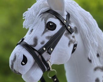 May include: A white plush hobby horse with grey spots, featuring a long white yarn mane, dark brown eyes, and a black faux leather bridle with silver-toned metal hardware. The horse head is crafted from soft fabric and includes a matching black rein.