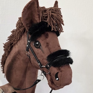 May include: A brown plush hobby horse with a dark brown yarn mane, black faux fur accents on the bridle, and a black leather-like bridle with silver-toned metal hardware. The horse features painted eyes and nostrils on a soft fabric surface.