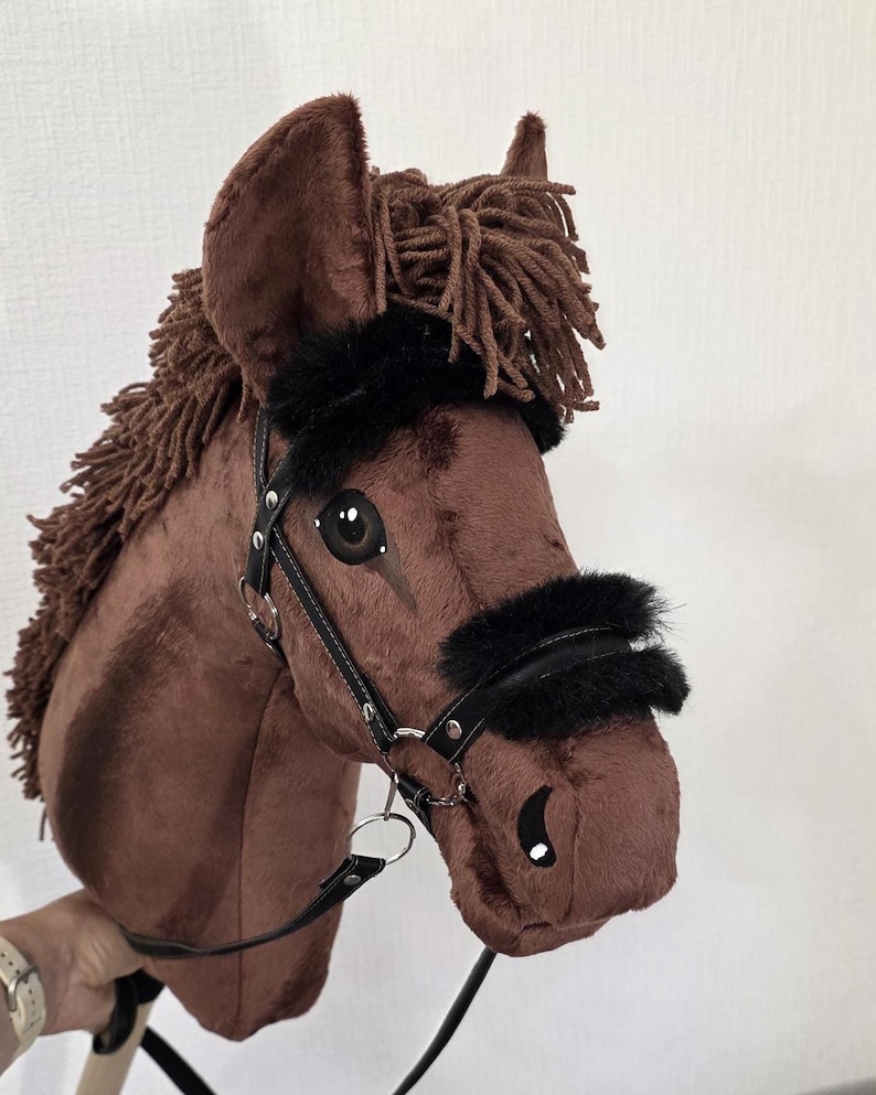 May include: A brown plush hobby horse with a dark brown yarn mane, black faux fur accents on the bridle, and a black leather-like bridle with silver-toned metal hardware. The horse features painted eyes and nostrils on a soft fabric surface.
