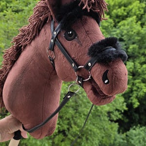 May include: A brown plush hobby horse toy featuring a dark brown yarn mane, black faux fur accents on the bridle, and a black leather-like halter with silver-toned metal rings and buckles, all mounted on a light-colored wooden stick handle.
