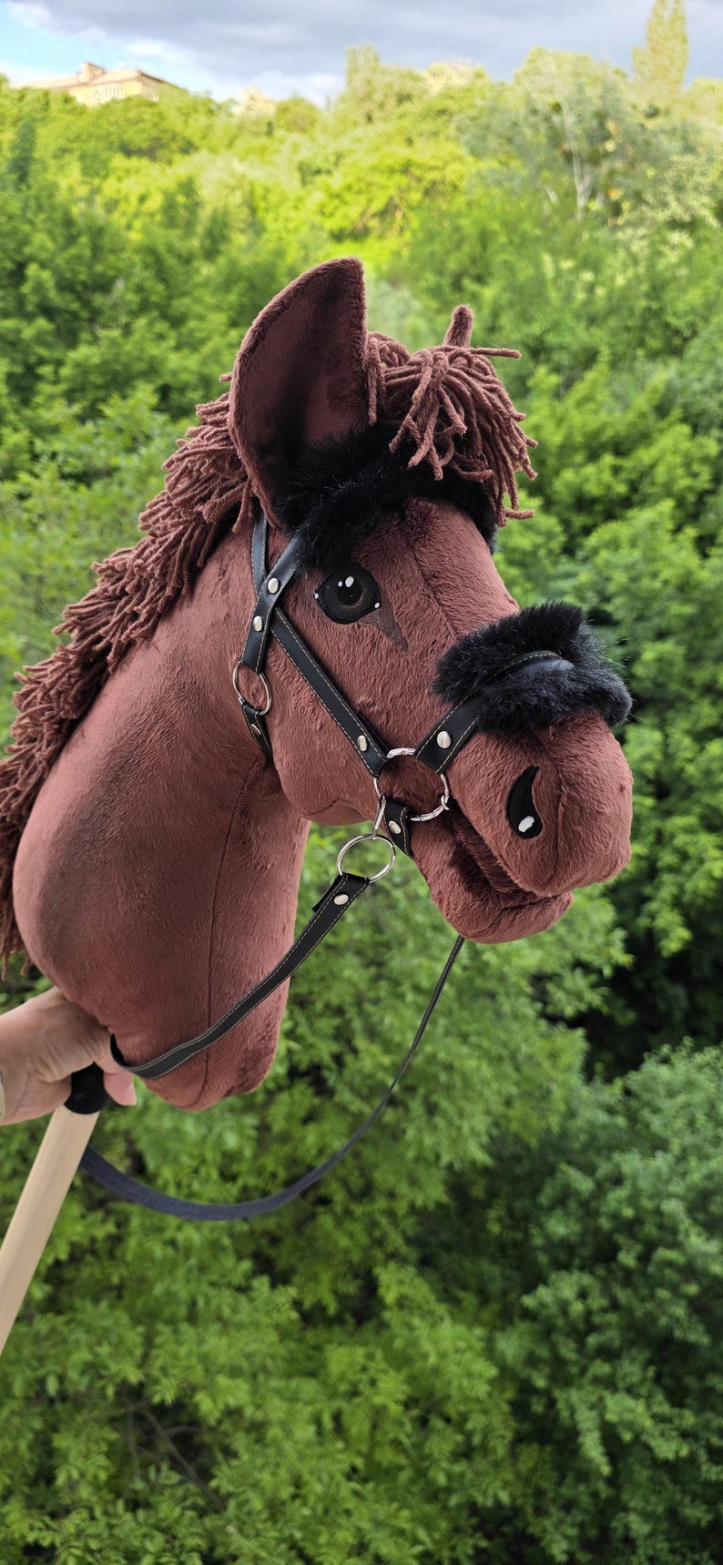 May include: A brown plush hobby horse toy featuring a dark brown yarn mane, black faux fur accents on the bridle, and a black leather-like halter with silver-toned metal rings and buckles, all mounted on a light-colored wooden stick handle.