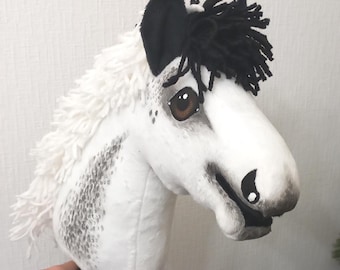 May include: A white and black hobby horse head with a black mane and ears. The horse has brown eyes and gray spots on its face and neck. The horse is held by a black handle.