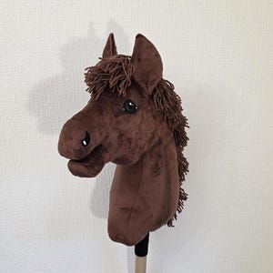 May include: A brown plush hobby horse with a soft velvet-like texture, featuring a dark brown yarn mane and forelock, black and white stitched eyes, and a black nostril detail, all mounted on a sturdy wooden stick handle for play.
