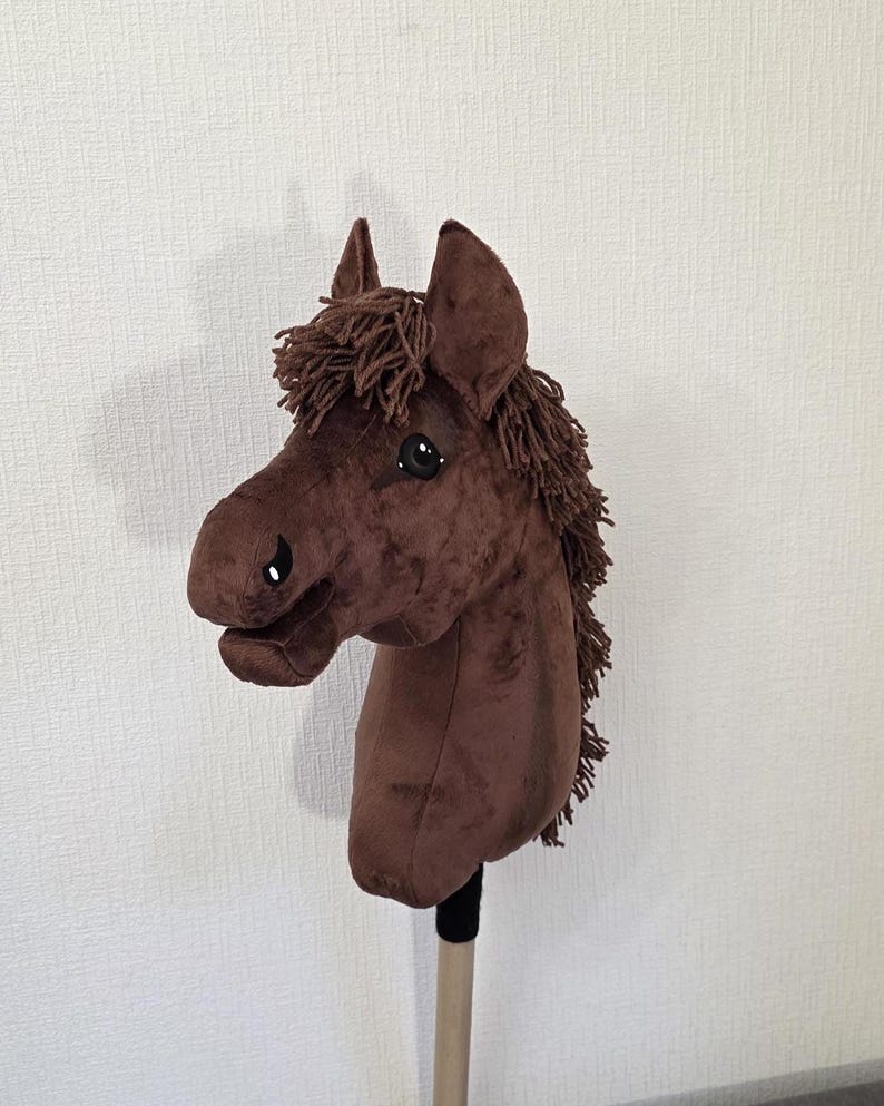 May include: A brown plush hobby horse with a soft velvet-like texture, featuring a dark brown yarn mane and forelock, black and white stitched eyes, and a black nostril detail, all mounted on a sturdy wooden stick handle for play.