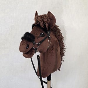 May include: A brown plush hobby horse with a dark brown yarn mane and forelock. The horse features a black faux leather bridle with silver-toned metal rings and black faux fur padding on the noseband and browband, mounted on a wooden stick.