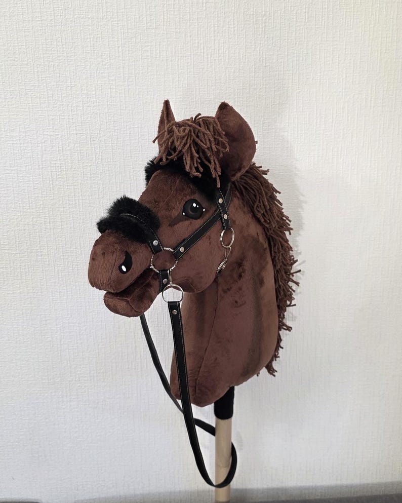 May include: A brown plush hobby horse with a dark brown yarn mane and forelock. The horse features a black faux leather bridle with silver-toned metal rings and black faux fur padding on the noseband and browband, mounted on a wooden stick.
