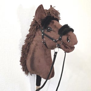 May include: A brown plush hobby horse with a dark brown yarn mane and forelock. The horse features a black faux leather bridle with silver-tone metal rings and black faux fur trim on the noseband and browband. It is attached to a wooden stick.