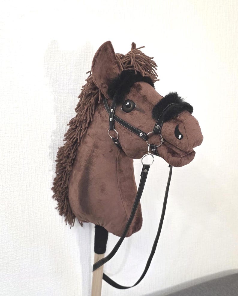 May include: A brown plush hobby horse with a dark brown yarn mane and forelock. The horse features a black faux leather bridle with silver-tone metal rings and black faux fur trim on the noseband and browband. It is attached to a wooden stick.