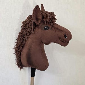May include: A brown plush hobby horse with a soft velvet-like texture, featuring a dark brown yarn mane and forelock, black plastic eyes, and stitched nostril details. The horse head is mounted on a wooden stick with a black fabric handle wrap.