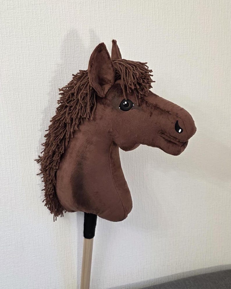 May include: A brown plush hobby horse with a soft velvet-like texture, featuring a dark brown yarn mane and forelock, black plastic eyes, and stitched nostril details. The horse head is mounted on a wooden stick with a black fabric handle wrap.
