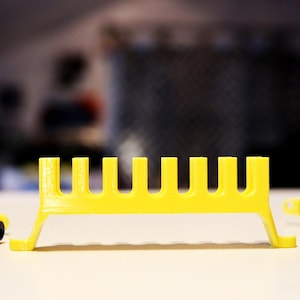 May include: A bright yellow plastic stand featuring a horizontal bar with eight evenly spaced vertical slots. The structure is supported by two angled legs on either side, creating a raised platform design with a smooth, matte finish.