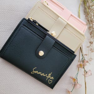 May include: Three small wallets in black, beige, and pink. The black wallet has the name "Samantha" in gold script with a heart. Each wallet has a zippered compartment and a snap closure. The wallets are made of a smooth material.