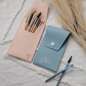 May include: Two makeup brush holders, one pink with the initials "SF" and a heart, and one blue with the name "Ellie" written in white. Both holders are made of faux leather and have a flap closure.