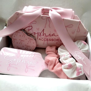 May include: A pink gift set containing a cosmetic bag, hairbrush, heart-shaped compact mirror, and scrunchies. The items are decorated with bow patterns and the name "Sophia". A pink ribbon with the name "Sophia" is tied around the set.