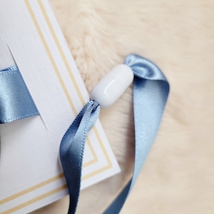 May include: A white ceramic bead with a blue ribbon attached to a white card with gold trim.