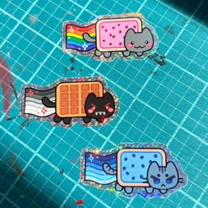 May include: Three holographic stickers featuring cat-like creatures with food bodies. The top cat has a pink pastry body and rainbow trail. The middle cat has a waffle body and grey-scale trail. The bottom cat has a blue pastry body and blue trail.
