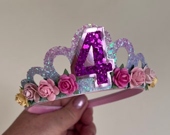 Puede incluir: Una diadema de purpurina rosa y morada con un gran número cuatro morado y flores rosas.