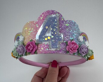 Puede incluir: Una corona brillante y colorida con el número 4 en el centro. La corona presenta purpurina rosa, morada y azul, con flores de papel y arcoíris. La corona está sujeta por una diadema rosa.