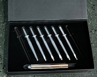 Op de afbeelding: Een set van zes zilveren en zwarte pennen en een stylus, gepresenteerd in een zwarte doos. De pennen hebben een strak, modern design en de doos heeft een getextureerde afwerking. De pennen zijn netjes gerangschikt in een met fluweel gevoerd compartiment.