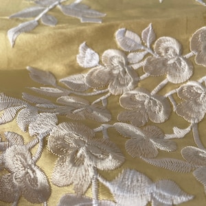 May include: Close-up of a yellow fabric with white embroidered floral designs. The fabric has a subtle sheen and the embroidery is detailed and intricate.