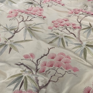 May include: A cream-coloured fabric with an embroidered floral pattern. The flowers are pink and the leaves are green and blue.