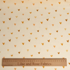 May include: Cream colored fabric with a repeating pattern of small brown and orange hearts. A wooden ruler with the text "Lilac and Rose Fabrics" is at the bottom of the image.