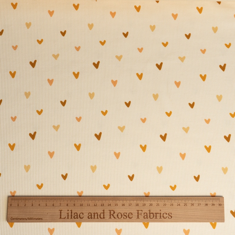 May include: Cream colored fabric with a repeating pattern of small brown and orange hearts. A wooden ruler with the text "Lilac and Rose Fabrics" is at the bottom of the image.