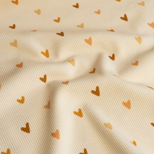 May include: A close-up of a cream-colored fabric with a subtle ribbed texture. The fabric is covered in a repeating pattern of small brown and orange hearts.