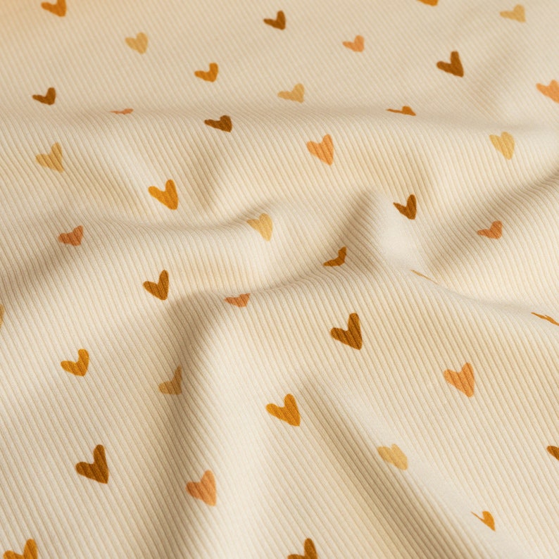 May include: A close-up of a cream-colored fabric with a subtle ribbed texture. The fabric is covered in a repeating pattern of small brown and orange hearts.