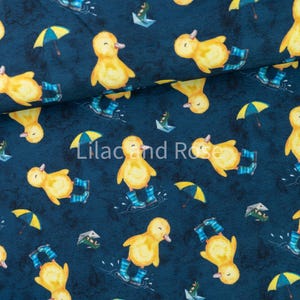 May include: Dark blue fabric featuring a pattern of yellow ducklings in blue wellington boots, yellow umbrellas, and paper boats. The text "Lilac and Rose" is printed on the fabric.