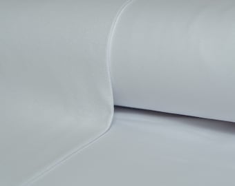 May include: A close-up of a white fabric, possibly a sheet or textile, with a smooth, slightly textured surface. The fabric is folded over, revealing a rolled edge. The color is a soft, light white.