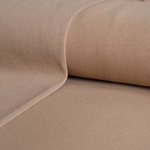 May include: Close-up of a roll of tan fabric. The fabric has a ribbed texture and a smooth, folded edge. The colour is a warm, neutral tone, suitable for various sewing and crafting projects. The fabric appears soft and pliable.