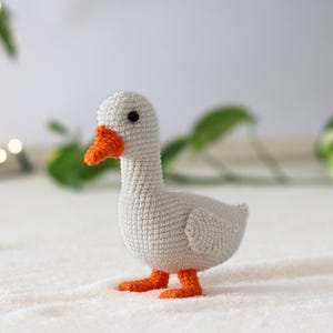 Puede incluir: Un ganso blanco de crochet con pico y patas naranjas. El ganso est&aacute; de pie sobre una superficie blanca.